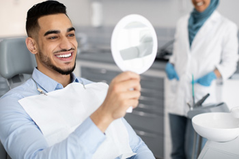 Man in dental chair smiling at his reflection in handheld mirror