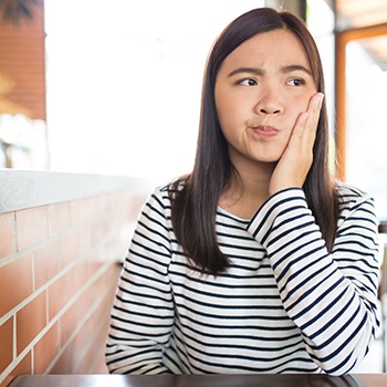 Woman in striped shirt sitting at table rubbing jaw in pain